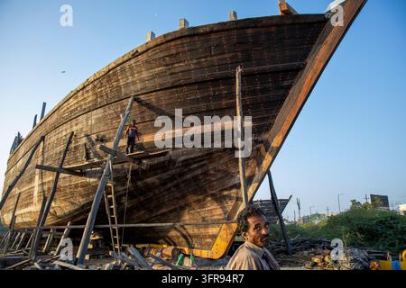 Uomo in primo piano in primo piano si trova vicino allo scafo di una nave gigante in legno con un operaio in equilibrio su una piattaforma di legno, Gujarat, Mandvi, India Foto Stock