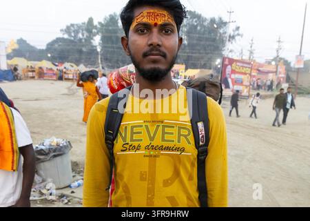 Giovane uomo con un tilak Mahakal sulla fronte e una camicia gialla si trova nel campo affollato del Maha Kumbh Mela, Uttar Pradesh, Prayagraj, India Foto Stock