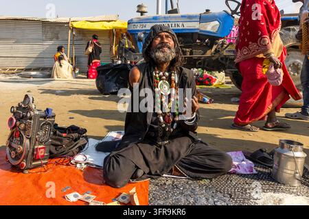 Uomo barbuto in nero con perline seduto per terra che mendica accanto a un trattore al Maha Kumbh Mela, Uttar Pradesh, Prayagraj, India Foto Stock