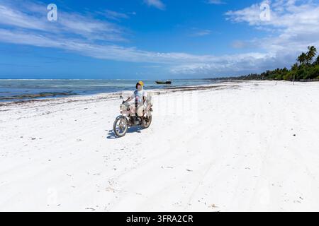 Uomo con cappello kofia giallo in sella a una moto rossa lungo una vasta spiaggia di sabbia bianca con un laghetto turchese e una barca di legno sotto un cielo blu profondo Foto Stock