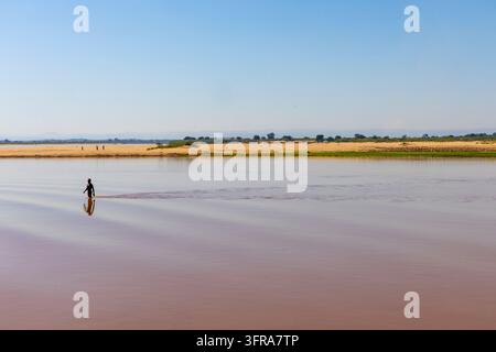 Un uomo solitario avanza nell'acqua calma del fiume sotto lunghe ombre di alberi, Menabe, Tsiribihina, Madagascar Foto Stock