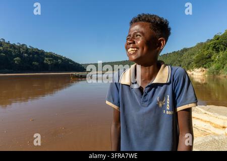 Un giovane uomo sorridente ride accanto al fiume mentre una barca naviga oltre colline boscose, Menabe, Tsiribihina, Madagascar Foto Stock
