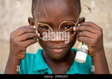 Un ragazzo sorridente indossa un paio di occhiali fatti a mano con filo metallico e tiene una banconota arrotolata infilata in una bottiglia, Regione Marittima, Lomé, Togo Foto Stock