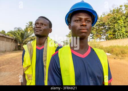 Un ritratto ravvicinato mostra due operai edili con giubbotti di sicurezza gialli, uno con un casco blu, Regione Plateaux, Kpalime, Togo Foto Stock