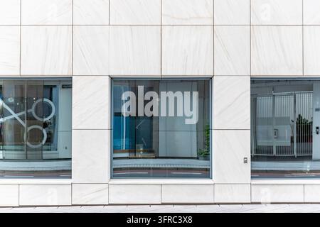 Facciata moderna in marmo bianco con tre grandi finestre in vetro, linee di pannelli geometriche pulite, riflessi sottili e un marciapiede urbano in primo piano. Foto Stock
