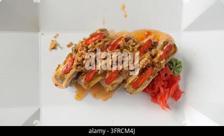 Rotoli di sushi tempura piccanti con cipolle fritte e chili sopra, serviti con zenzero e wasabi in una scatola da asporto Foto Stock