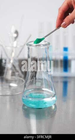 Mano di chimico che aggiunge una polvere chimica verde con una spatola in una beuta di Erlenmeyer contenente un liquido in un laboratorio moderno Foto Stock