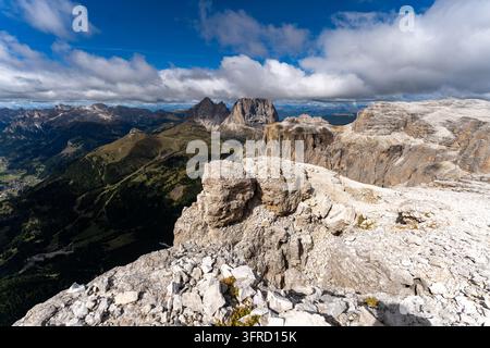 Una vista grandangolare da un alto altopiano roccioso rivela la drammatica catena montuosa delle Dolomiti sotto un cielo blu luminoso con nuvole sparse. Verde Foto Stock