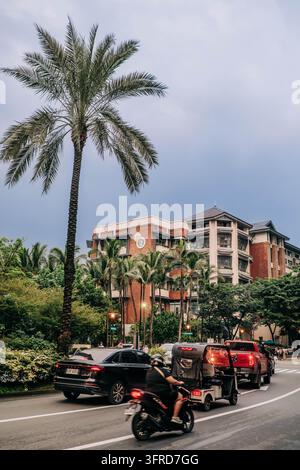 Manila, Filippine, 23 settembre 2026. Un motociclista e un triciclo motorizzato viaggiano lungo una strada cittadina accanto a macchine, palme e un edificio in mattoni rossi. Foto Stock