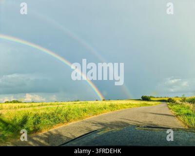 Vista panoramica di un doppio arcobaleno su un campo verde e una strada di campagna, vista da un'auto, dopo una pioggia estiva Foto Stock