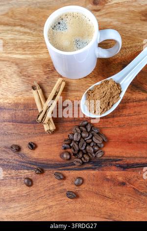 Caffè appena preparato in una tazza bianca, circondato da chicchi di caffè tostati, bastoncini di cannella aromatici e polvere di cannella su uno sfondo di legno Foto Stock