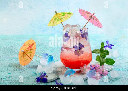 Un bicchiere di bevanda rinfrescante ghiacciata con fragole, limone e fiori viene servito con ombrelli di carta. Limonata fatta in casa alle bacche estive Foto Stock