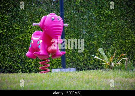Un giocattolo a molla a forma di elefante rosa acceso situato su un prato verde in un parco pubblico o in un parco giochi residenziale. Lo sfondo presenta una vegetazione curata Foto Stock