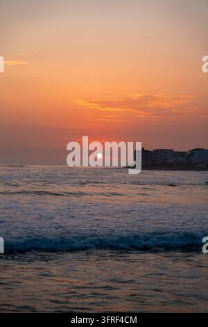 Tramonto arancione sulle onde dell'Oceano Pacifico alla spiaggia di San Bartolo a Lima, Perù, con edifici costieri silouettati all'orizzonte. Foto Stock