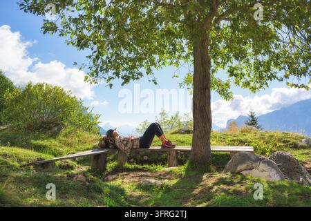 Escursionista donna sdraiata su una panca di legno che riposa sotto un albero verde Foto Stock