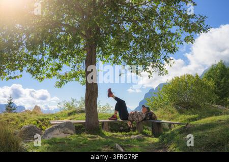 Escursionista donna sdraiata su una panca di legno che riposa sotto un albero verde Foto Stock