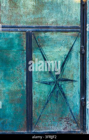 Primo piano di una vecchia porta metallica turchese con vernice arrugginita e screpolata e una stella di bussola blu scuro dipinta a mano Foto Stock