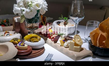 Tavola di salumi con cubetti di formaggio, carni stagionate, datteri e fiori commestibili, con olive, fiori bianchi e bicchieri di vino su un tavolo di ristorante a Turke Foto Stock