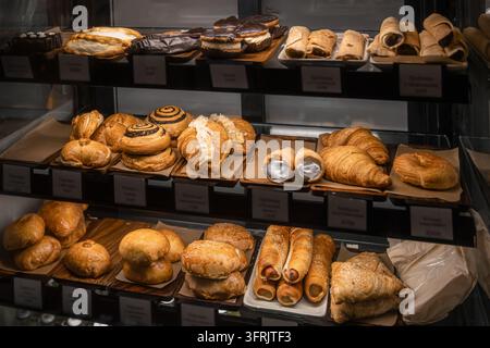 Ampia selezione di pasticcini appena sfornati esposti su scaffali all'interno di un bancone di panetteria in vetro. Croissant dorati, pasticcini sfogliati ripieni, rotoli al cioccolato Foto Stock
