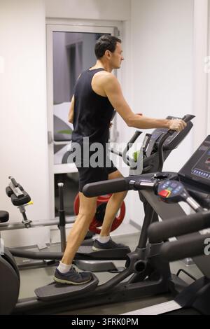 Uomo affascinante in nero si allena su una macchina da step in un moderno centro fitness - Uomo fa esercizio su un tapis roulant in una palestra. Indossa abbigliamento sportivo scuro. Sembra f Foto Stock