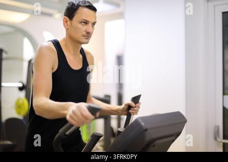 Uomo affascinante in nero si allena su una macchina da step in una palestra, primo piano – Uomo si allena su una bicicletta stazionaria in palestra. Espressione concentrata visibile durante Foto Stock