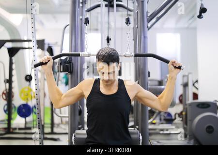 Uomo affascinante in nero si allena su una macchina per i pesi in una palestra vuota – L'uomo si allena in palestra usando una macchina a cavi. Indossa una canottiera nera. Concentrato su Foto Stock