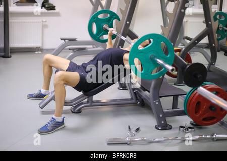 Uomo in pantaloncini neri si allena con bilanciere in una palestra vuota - L'uomo è sdraiato su un panca inclinata e preme il bilanciere. I piedi piantati saldamente sul pavimento per la stabilità Foto Stock