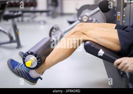 Uomo in nero si allena su macchina per le gambe in una palestra vuota – Persona esercita la gamba con una macchina da ginnastica. Piedi fissati su rullo imbottito. Scarpe, Foto Stock