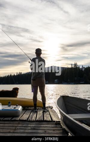Uomo che pesca su un molo di legno con il suo cane al tramonto Foto Stock