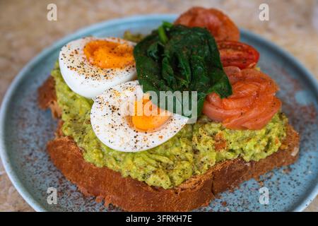 Una deliziosa fetta di toast artigianale è guarnita con avocado schiacciato, uova sode, rosette di salmone affumicato, spinaci e pomodori a fette. Foto Stock