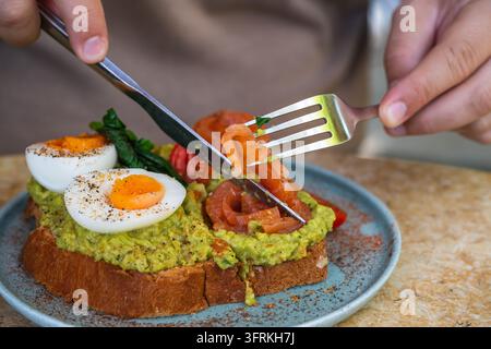 Un toast gourmet all'avocado con uova sode, salmone affumicato, pomodorini e spezie viene mangiato con coltello e forchetta su un piatto Foto Stock