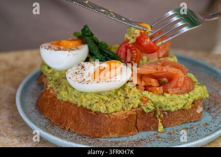 Un toast gourmet all'avocado con uova sode, salmone affumicato, pomodorini e spezie viene mangiato con coltello e forchetta su un piatto Foto Stock