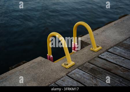 Scatto ad angolazione alta di una vivace scala metallica gialla su un molo di cemento che si estende in acque profonde e scure, enfatizzando l'accesso marittimo e le infrastrutture Foto Stock
