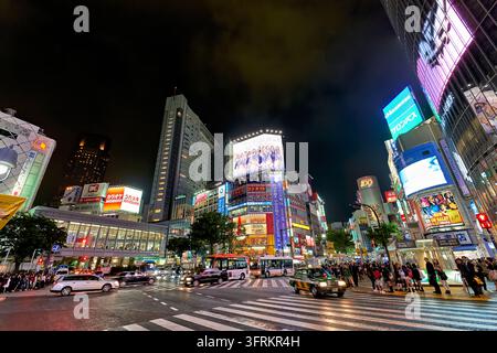 Taxi, autobus e pedoni al crocevia di Shibuya sotto cartelloni illuminati vicino alla stazione di Shibuya a Tokyo, Giappone. Foto Stock