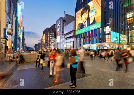 Pedoni in sfocatura di movimento attraversano una piazza sotto cartelloni pubblicitari nel quartiere di intrattenimento di Dotonbori a Namba, Osaka, Giappone, al crepuscolo Foto Stock