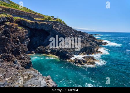 Grotta marina tra scogliere vulcaniche scure sopra un'insenatura turchese, con le onde dell'Atlantico che si infrangono sulle rocce di lava e sui muri della terrazza in pietra sul pendio soprastante Foto Stock