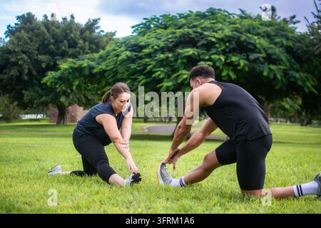 Due adulti in abbigliamento sportivo che eseguono esercizi di allungamento delle gambe insieme sull'erba di un parco pubblico, praticando l'allenamento di flessibilità e mobilità Foto Stock