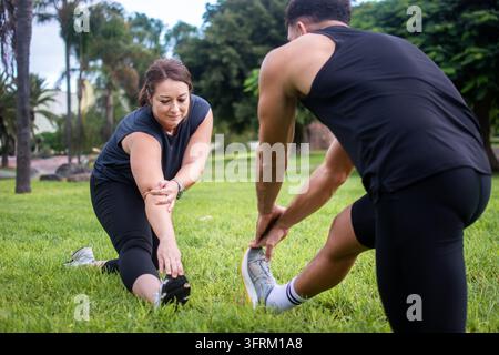 Due adulti in abbigliamento sportivo che eseguono esercizi di allungamento delle gambe insieme sull'erba di un parco pubblico, praticando l'allenamento di flessibilità e mobilità Foto Stock