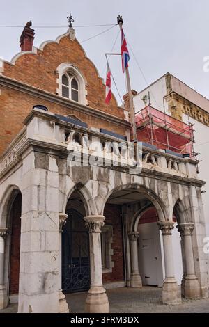 Città Vecchia, Gibilterra - 1 marzo 2026: Portico storico in pietra con archi e facciata di edificio in mattoni, bandiere e ponteggi sotto un cielo nuvoloso. Patrimonio architettonico e meta turistica nel Foto Stock
