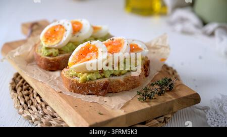 Due fette di pane a lievitazione naturale croccante ricoperte di avocado frantumato e uova sode morbide e vivaci. La luce naturale soffusa del mattino mette in risalto il tessuto cremoso Foto Stock