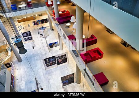 Vista ad angolazione alta di pannelli di esposizione fotografica su un pavimento di marmo bianco in un atrio a più livelli di un edificio pubblico con sedute lounge rosse, Atene, Grecia. Foto Stock