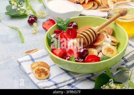 Piccoli pancake con salsa di panna e miele, fragole e ciliegie serviti con menta fresca in una ciotola di ceramica, messa a fuoco selettiva. Mini pancake alla moda Foto Stock