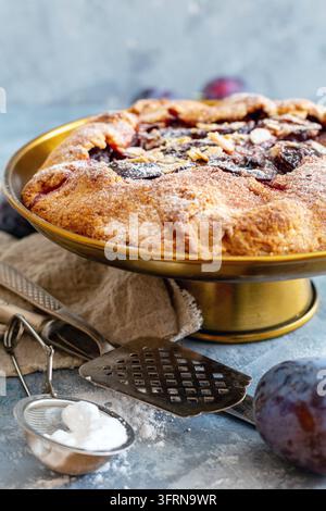 Supporto per torte in bronzo con ripieno di prugne e scaglie di mandorle e prugne blu fresche su un tavolo di cemento strutturato Foto Stock