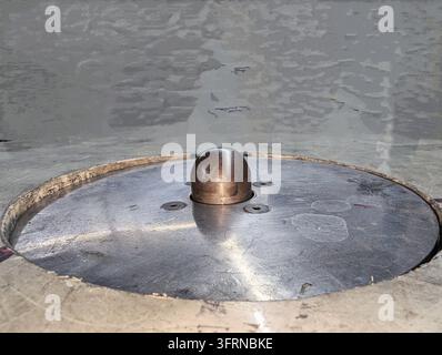 Primo piano di una piastra metallica circolare con pulsante centrale in metallo arrotondato e tre piccoli fori per viti, superficie graffiata e consumata che riflette la luce dall'alto, co Foto Stock