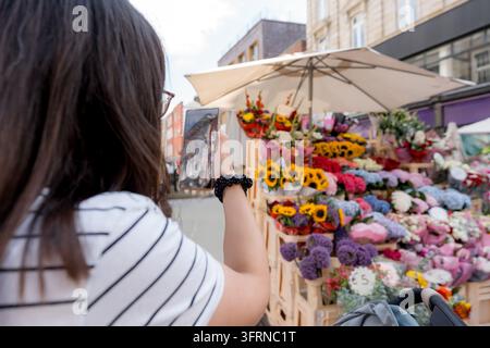 Donna usa uno smartphone per fotografare bouquet colorati a una bancarella di fiori all'aperto su Grafton Street, catturando una vivace scena di shopping nel centro di Dub Foto Stock