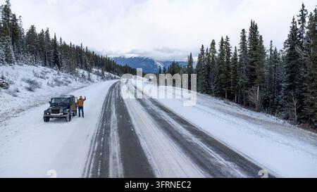 Vista aerea con drone a bassa quota di un giovane uomo felice che fa il segno della pace accanto a un veicolo fuoristrada nero su un'autostrada innevata in Alberta, Canada, incorniciato da Foto Stock