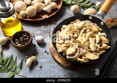 Vista aerea rustica di funghi saltati e cipolla affettata in una padella nera, circondati da olio d'oliva, aglio, grani di pepe e funghi freschi su un d Foto Stock