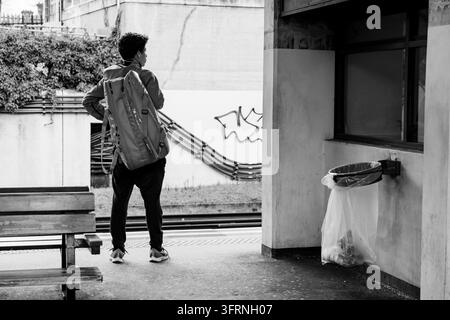 UOMO CON ZAINO IN ATTESA SU UN BINARIO, ARCHIVIO REPORTAGE DELLA METRO DI LONDRA, LONDRA, INGHILTERRA, 2019: Un giovane con uno zaino è visto di spalle mentre aspetta su un binario accanto a un cestino in questa scena in bianco e nero. INFO: La metropolitana di Londra, affettuosamente conosciuta come “la Tube”, è la prima ferrovia sotterranea passeggeri del mondo, aperta nel 1863. Con 11 linee e 272 stazioni, questa rete iconica trasporta milioni di pendolari e visitatori ogni giorno. Foto Stock