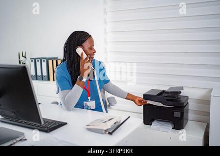 Professionista amministrativa al telefono mentre utilizza una stampante in ufficio Foto Stock