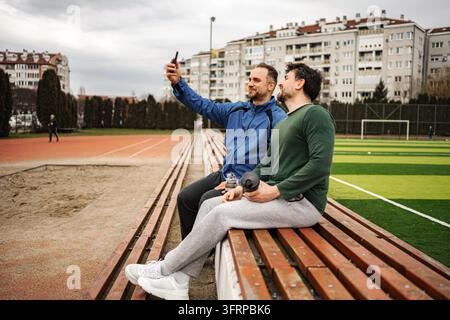 Amici maschi seduti sulle panchine dello stadio a fare selfie Foto Stock